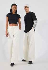 Un haut noir court et un pantalon blanc large sur un modèle, associés à des chaussures noires. L'homme porte une chemise noire avec des graphiques beiges sur les manches.