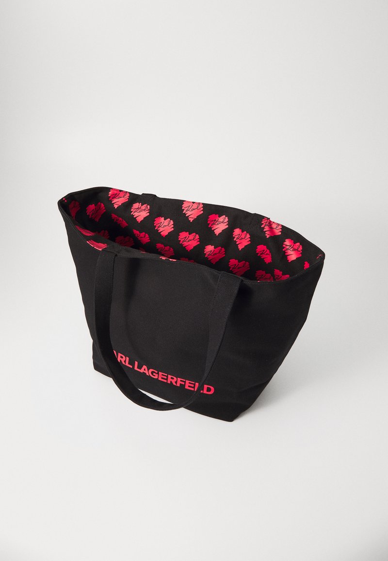 Sac en toile noir avec le logo "Karl Lagerfeld" en rose. L'intérieur présente un motif de cœurs rouges sur fond noir. Deux poignées robustes.