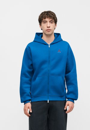 Jeune homme aux cheveux bouclés portant un sweat à capuche bleu zippé avec un logo rouge sur la poitrine, debout devant un fond uni.