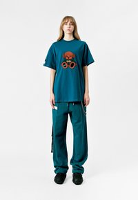 Camiseta de gran tamaño en color azul verdoso con un gráfico texturizado de un oso marrón, combinada con pantalones azul verdoso y zapatos negros texturizados. Diseño simple y funcional.