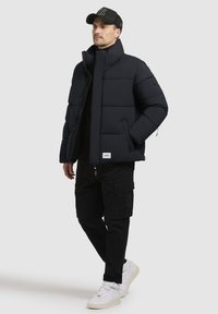 Tumesinine puffermantel kõrge kaelusega, mida on kombineeritud mustade cargo-pükstega ja valgete spordijalatsitega. Omab lukku ja nööpe, samuti plaadi detaili.