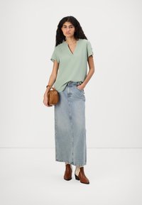 Top verde chiaro a maniche corte con scollatura a V, abbinato a una lunga gonna di jeans. Una borsa marrone e stivaletti alla caviglia completano il look, evidenziando uno stile casual.