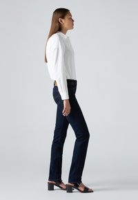 Γυναίκα με μακριά καστανά μαλλιά που φοράει άσπρο μπλουζάκι, σκούρα μπλε τζιν και μαύρες γόβες με τακούνι, στέκεται προφίλ σε γκρι φόντο.