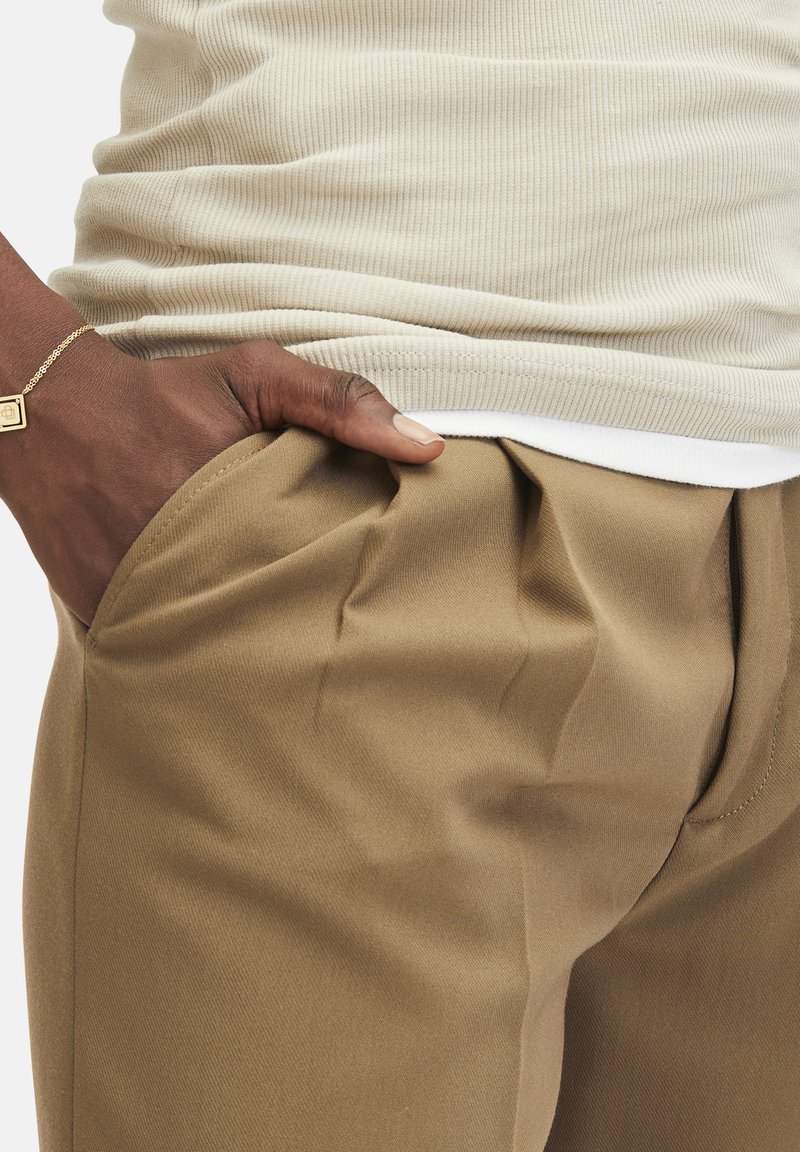 Beige geribde tanktop gecombineerd met lichtbruine broeken, met zichtbare zakken en zachte stof met lichte plooien bij de tailleband.