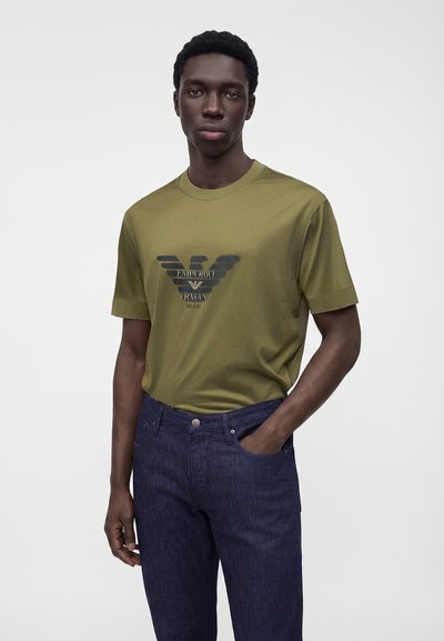 Άνδρας που φοράει ελιάς πράσινη μπλούζα Emporio Armani και σκούρα μπλε τζιν, στέκεται με το ένα χέρι στην τσέπη σε απλό φόντο.