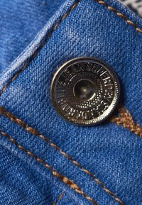 Tela de mezclilla azul con un primer plano de un botón de metal plateado que presenta un grabado y un agujero central, rodeado de costuras doradas.