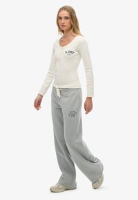 Joven mujer caminando, vistiendo una blusa de manga larga color crema con botones y pantalones de chándal de pierna ancha grises, con zapatillas beige.