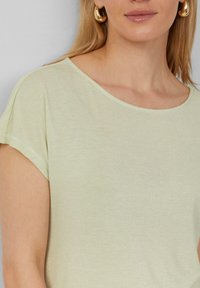 Hellegrünes Top mit Rundhalsausschnitt und kurzen Ärmeln, das eine subtile glänzende Optik und eine feine Textur aufweist. Im Bild sind goldene Creolen zu sehen.