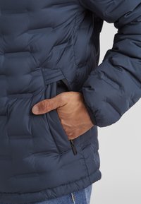 Dunkelblaue Puffjacke mit gestepptem Design, einem seitlichen Reißverschlussfach und elastischen Bündchen, die einen gemütlichen und isolierten Sitz bieten.