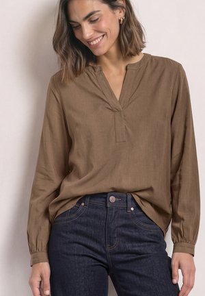 Femme souriante en regardant vers le bas, portant un chemisier marron à manches longues et col en V, rentré dans un jean bleu foncé taille haute sur un fond uni.