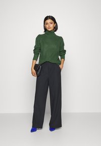 Zeleni rebrasti turtleneck pulover, črne široke hlače in modri čevlji s koničastim nosom; model drži črno torbico.