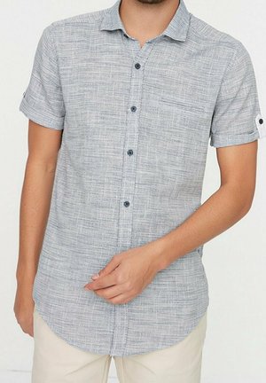 Uomo che indossa una camicia a maniche corte grigio chiaro con bottoni neri e pantaloni beige, mano che sistema la parte frontale della camicia.