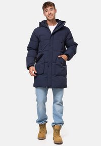 Geacă puffer bleumarin cu glugă, design matlasat, închidere cu nasture și fermoar, și două buzunare frontale, purtată cu blugi albaștri deschiși și cizme bej.