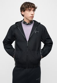 Νεαρός άνδρας που φοράει μαύρο ζακέτα με κουκούλα και φερμουάρ New Balance πάνω από μωβ φούτερ και καρό πουκάμισο, κοιτάζει προς τα δεξιά με τα χέρια στις τσέπες.