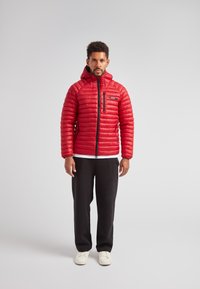 Giacca puffa rossa con texture trapuntata, cappuccio e zip frontale. Abbinata a pantaloni neri e sneakers bianche. Vista frontale su uno sfondo neutro.