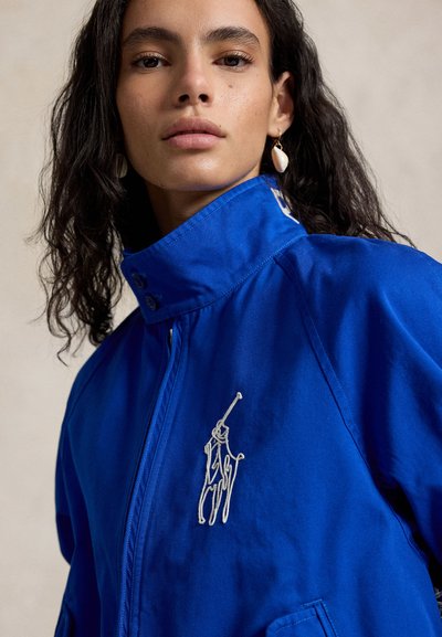 Jonge vrouw met lang donker haar draagt een blauwe jas met hoge kraag en een wit geborduurd logo van een paard en ruiter, plus een schelpenoorbel.