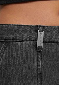 Jean en denim gris foncé avec une surface texturée, doté d'une fermeture éclair en métal et d'une étiquette indiquant "2Y Studios" près de la taille.