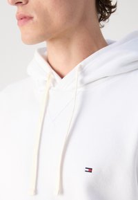 Weißer Kapuzenpullover mit Kordelzug, versehen mit einem kleinen rot-, weiß- und blaufarbenen Logo auf der Brust. Weicher Stoff und klassischer Schnitt.