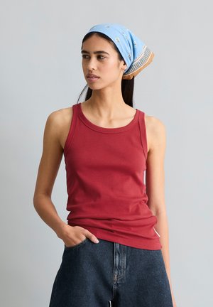 Frau trägt ein rotes ärmelloses Oberteil, blaues florales Kopftuch und dunkle Jeans mit einer Hand in der Tasche, blickt seitlich vor schlichtem Hintergrund.