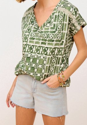 Grünes gemustertes Oberteil mit V-Ausschnitt und kurzen Ärmeln, kombiniert mit hellen Denim-Shorts mit ausgefranselten Kanten. Als Accessoires werden mehrere Armbänder getragen.