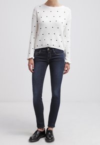 Suéter blanco de canalé con lunares negros, combinado con jeans ajustados azul oscuro y zapatos negros de cordones. Atuendo simple y casual.