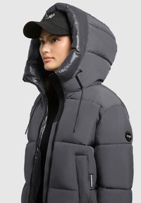 Pelēks pūkains apģērbs ar lielu kapuci, rāvējslēdzēja aizdari, elastīgām auklām un mīkstu tekstūru. Uz piedurknes ir logo uzlīme.