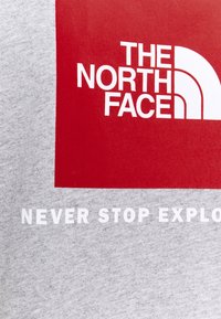 Červený čtverec s bílým logem "The North Face" na šedém textilním pozadí s částečným textem "Nikdy nepřestávejte objevovat."