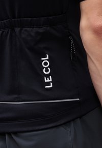 Tuvplāns melnai sporta jaciņas kabatai ar baltu izšuvumu “LE COL” logotipu un daļēji redzama roka melnos un pelēkos apģērbos.