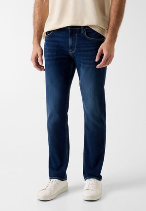 Mann trägt dunkelblaue Jeans mit geradem Bein und weiße Sneakers, steht vor einem schlichten hellen Hintergrund.