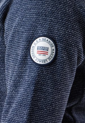 Marineblåt tekstureret stof med et rundt logo-patch med "Seaside Nautical Legacy" og røde striber. Materialet fremstår holdbart og indbydende.