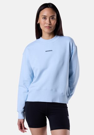 Jonge vrouw draagt een lichtblauwe Rossignol-trui en zwarte korte broek, staand tegen een effen lichte achtergrond.