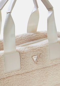 Be�żowa torba tote z sztucznego owczej wełny z kremowymi uchwytami, złotym logo oraz zamknięciem na zamek. Miękka faktura z wyraźnie zarysowanym kształtem.