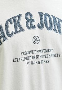 T-shirt en coton de couleur claire avec un texte bleu foncé indiquant "JACK & JONES" ainsi que des détails supplémentaires sur la création de la marque.