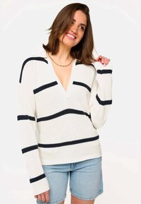 Pull en tricot crème avec des rayures horizontales bleu marine, possède un col en V et des manches longues, conçu pour une coupe décontractée. Associé avec un short en jean.