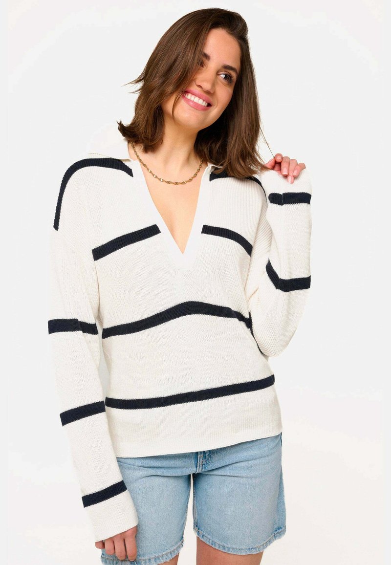 Pull en tricot crème avec des rayures horizontales bleu marine, possède un col en V et des manches longues, conçu pour une coupe décontractée. Associé avec un short en jean.