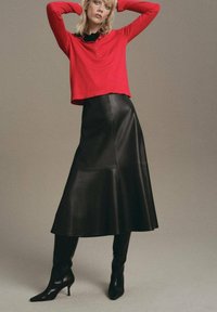 Haut rouge à manches longues avec des boutons, associé à une jupe midi en cuir noir et des bottes noires à talons pointus au genou, sur fond neutre.