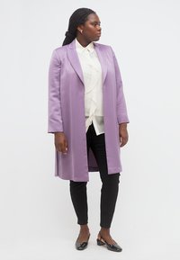 Γυναίκα με πλεξούδες στα μαλλιά, ντυμένη με λιλά παλτό, λευκό πουκάμισο με βολάν, μαύρο παντελόνι και μαύρες γόβες, στέκεται μπροστά σε λευκό φόντο.