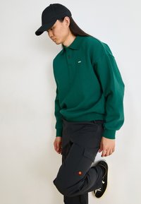 Groene sweatshirt met een kraag en twee knopen, gecombineerd met zwarte cargo broek met een klein gekleurd logo. Zwarte pet en schoenen.