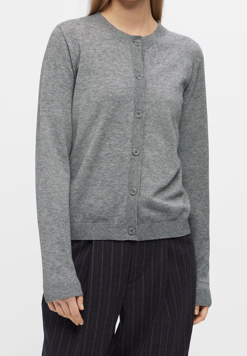 Femme portant un cardigan gris à boutons avec des manches longues et un pantalon à fines rayures foncées, debout devant un fond blanc.