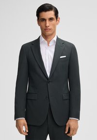 Giacca di abito grigio scuro con un design semplice, due bottoni e colletto a punta. Camicia bianca sotto, con dettaglio della tasca sul petto.