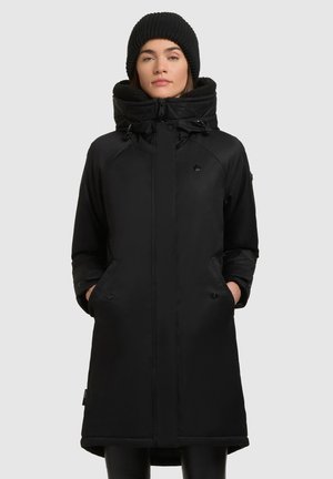 Vrouw in een zwarte winterjas met kap en zakken, en een zwarte gebreide muts, staande tegen een effen lichtgrijze achtergrond.