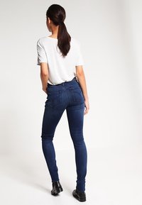 Jean slim fit bleu foncé à taille haute, avec poches arrière et texture lisse. Assorti à une chemise à manches courtes gris clair.