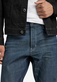 Czarna kurtka dżinsowa z zapięciem na guziki, noszona na niebieskich dżinsach z kontrastowymi przeszyciami i dopasowanym krojem.