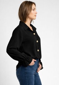 Mujer con blusa negra con botones dorados y vaqueros azules, de perfil con las manos en los bolsillos contra un fondo blanco liso.