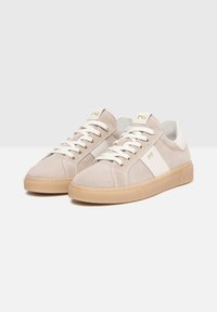 Beige suede sneakers with white accents and laces, featuring a textured upper, tan rubber sole, and gold logo detail on the tongue.