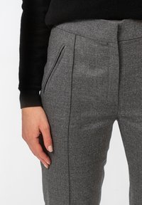 Szare, dopasowane spodnie o gładkiej fakturze, z płaskim przodem i kieszeniami bocznymi, noszone z czarnym swetrem. Zbliżenie na talia i dłoń.