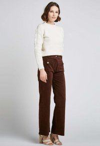 Pull crème avec des détails texturés sur les manches, associé à un pantalon en velours côtelé marron avec des poches avant et des boutons blancs. Sandales à talons beiges.