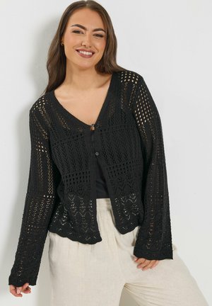 Lächelnde Frau, die eine schwarze, gehäkelte Strickjacke über einem schwarzen Oberteil und hellbeigen Hosen trägt, posiert vor einem weißen Hintergrund.