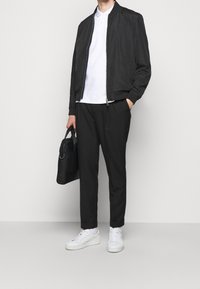 Homme portant une veste bomber noire, une chemise polo blanche, un pantalon noir, des baskets blanches, tenant une mallette noire dans la main gauche.