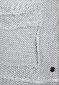 Grå og hvidt vævet stof med et struktureret mønster, der har en lomme og et rundt logo med sort finish. Kanterne har en udfrafrayed detalje.